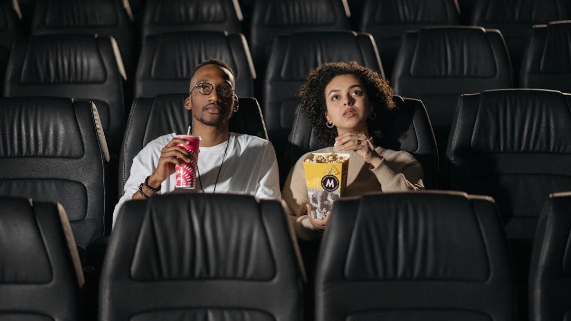 A couple sits in a cinema, enjoying popcorn while watching a movie.